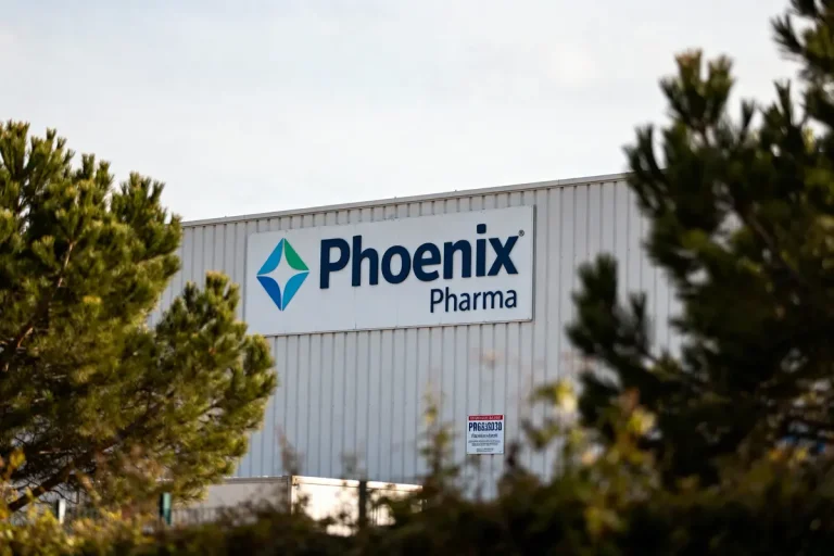 Bâtiment industriel avec logo Phoenix Pharma visible au milieu d'arbres verdoyants. Luminosité extérieure naturelle.