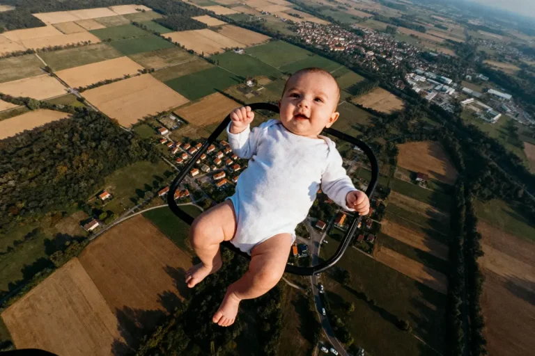 Bébé en grenouillère blanche dans un harnais au-dessus de paysages agricoles depuis une grande hauteur, ciel dégagé.