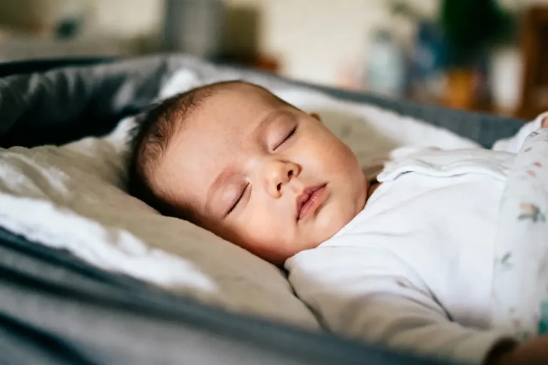 Bébé endormi paisiblement dans un lit, vêtu de blanc, couvert d'une couverture colorée.