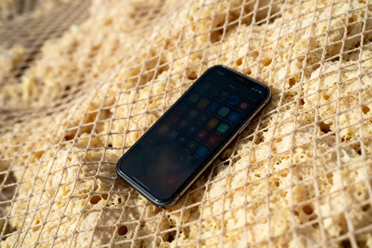 Smartphone noir sur fond de structure grillagée, surface beige poreuse en arrière-plan.