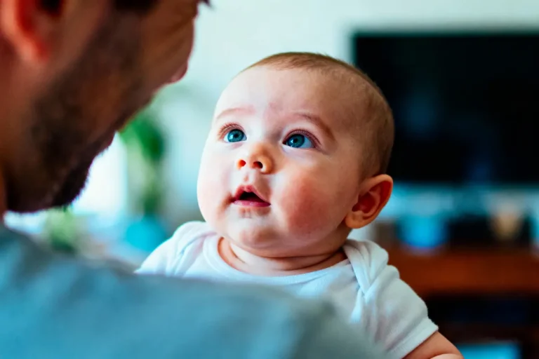 Un bébé aux yeux bleus regarde vers un adulte, fond flou en intérieur.