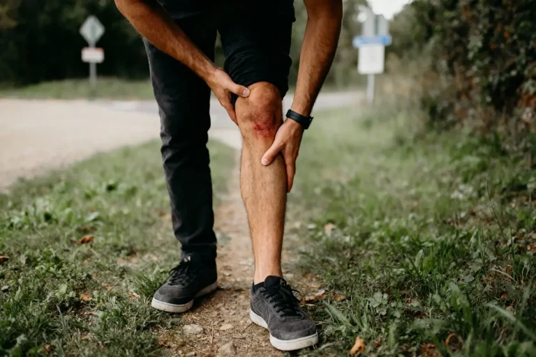 Un homme avec entorse au genou gauche sur un chemin, examine sa blessure, portant un pantalon noir.
