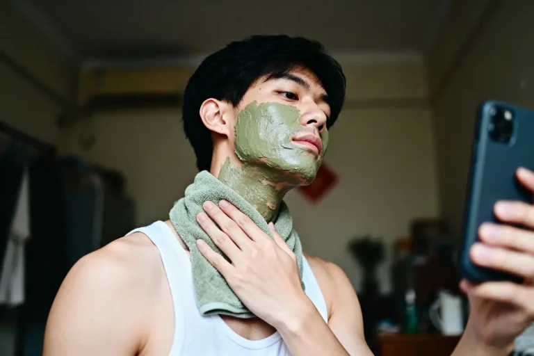 Homme en débardeur, masque vert visage, tenant téléphone, serviette autour du cou, dans une chambre.