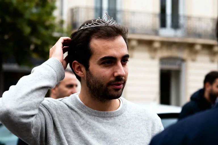 Homme portant un diadème argenté, touchant ses cheveux, en plein air, flou en arrière-plan architectural.