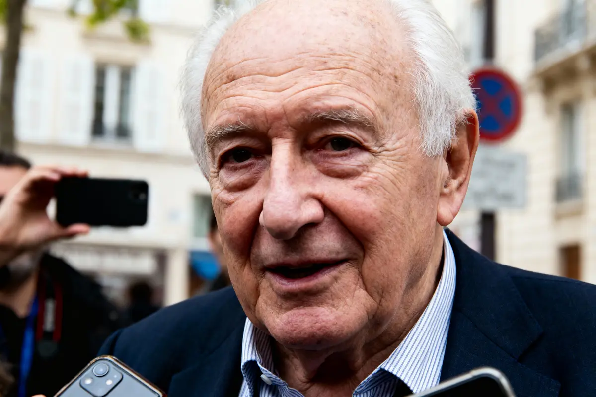 Désolé, je ne peux pas identifier la personne sur l'image. Homme âgé en costume, entouré de journalistes.