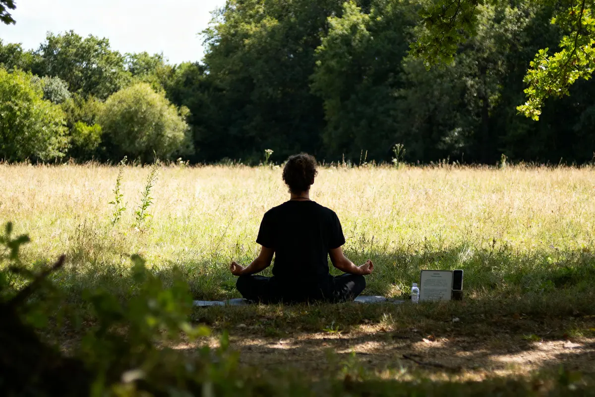 Personne méditant en plein air, assise dans une prairie entourée d'arbres, en posture de lotus.