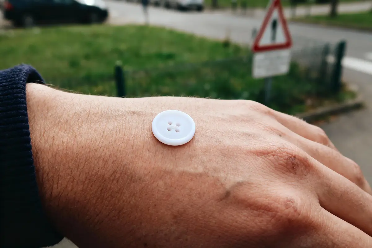 Bouton blanc posé sur une main extérieur, fond flou avec route verte et panneau.