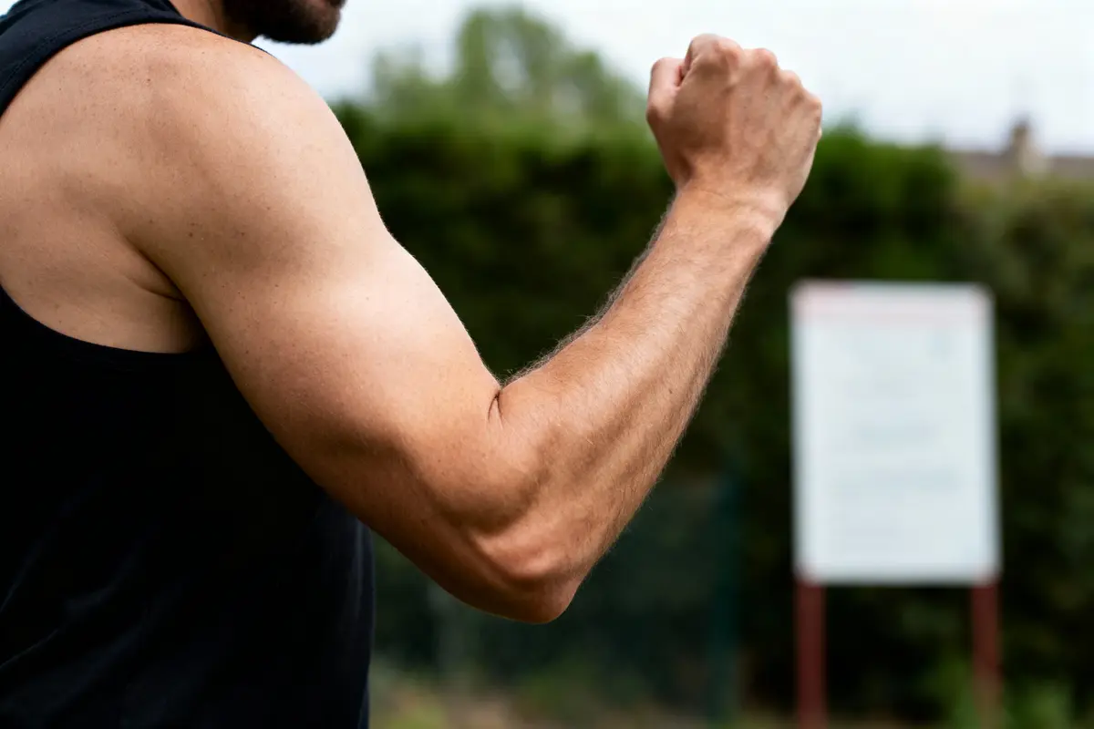 Bras musclé d'un homme en débardeur noir, poing serré, fond flou de verdure.