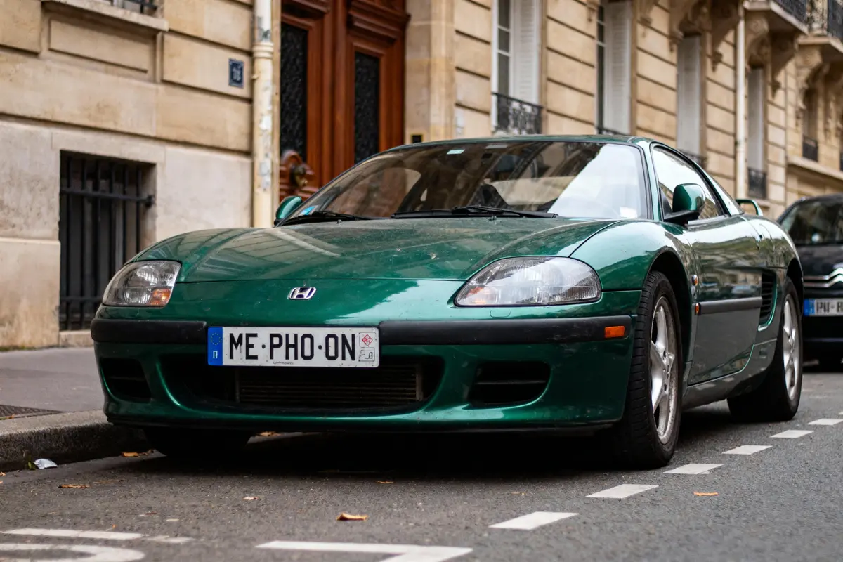Voiture verte stationnée devant un bâtiment en pierre avec plaque immatriculation humoristique.