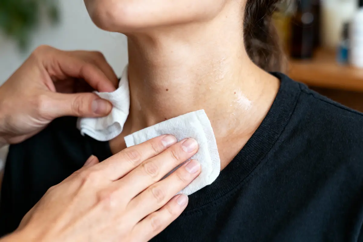 Deux mains nettoyant cou d'une personne avec des lingettes blanches, portant un t-shirt noir.