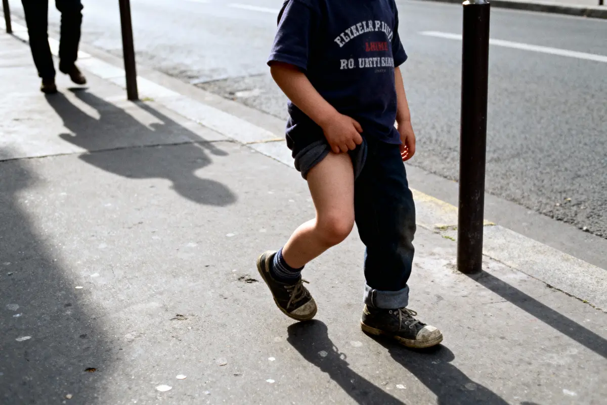 Enfant marchant sur un trottoir, pantalon relevé, ombre projetée au sol, lumière du jour.
