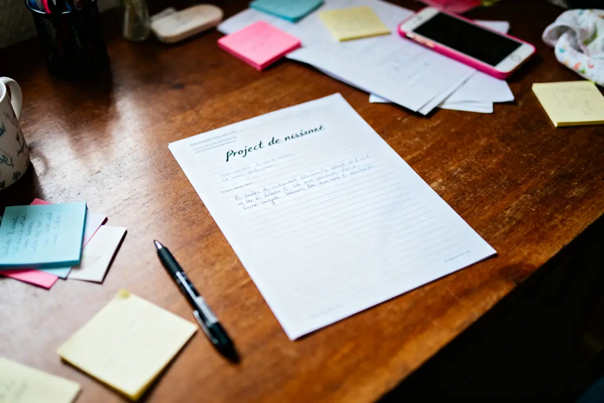 Feuille de projet manuscrite sur table en bois, avec stylos et notes adhésives colorées.