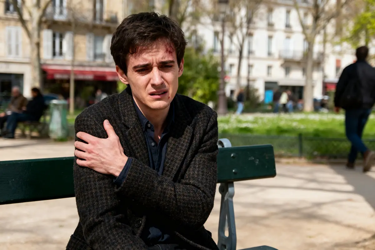 Homme assis sur un banc, se tenant l'épaule avec une expression douloureuse, parc en arrière-plan.