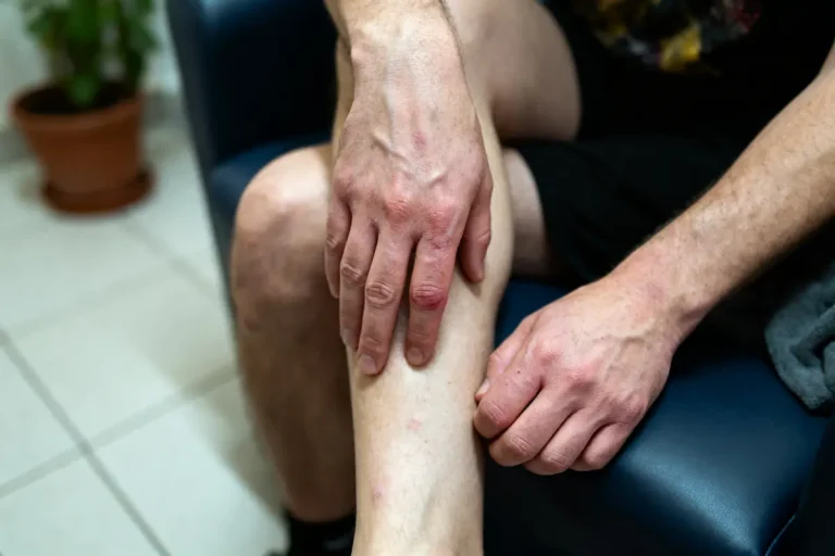 Un homme assis touche sa jambe, posée sur une chaise bleue, lumière intérieure.