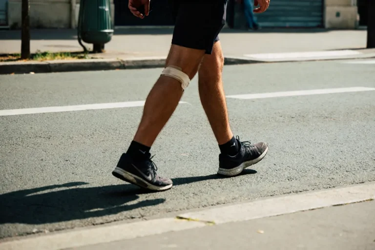 Personne marchant sur un trottoir, genou bandé, portant chaussures de sport et short, jour ensoleillé.